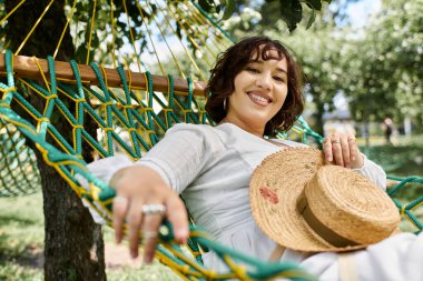 Beyaz elbiseli ve hasır şapkalı genç bir kadın yemyeşil bir hamakta dinleniyor..
