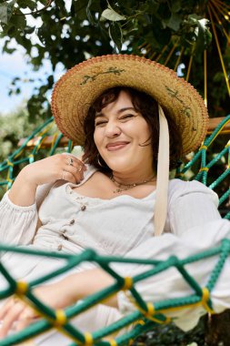 A young woman in a white dress and straw hat relaxes in a hammock in a lush summer garden. clipart