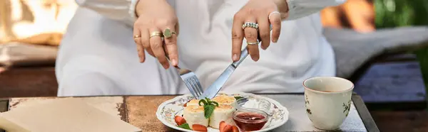 stock image A young woman in a white dress enjoys a summer brunch outdoors, cutting into a savory dish with a knife and fork.