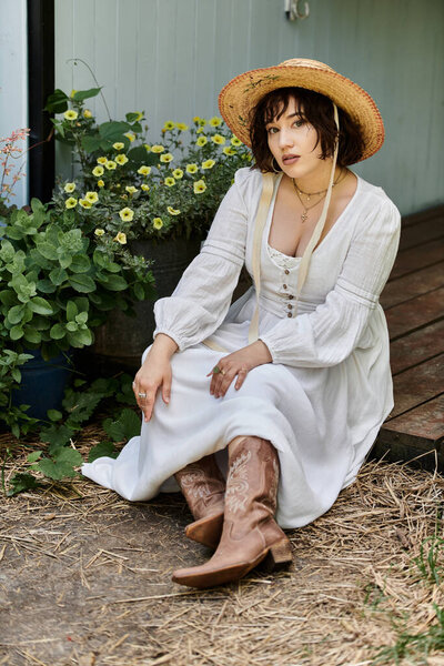 Молодая женщина в белом платье и соломенной шляпе сидит в летнем саду в окружении зелени.