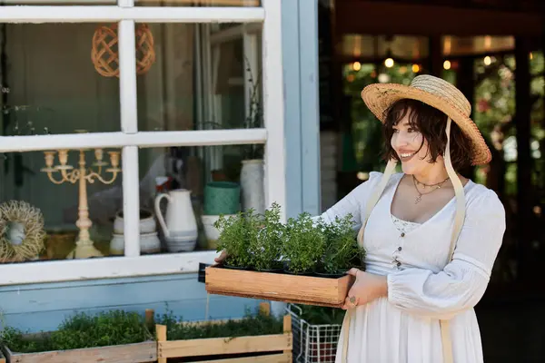 stock image A woman in a white dress and straw hat smiles while carrying a tray of potted plants in a sunny summer garden.
