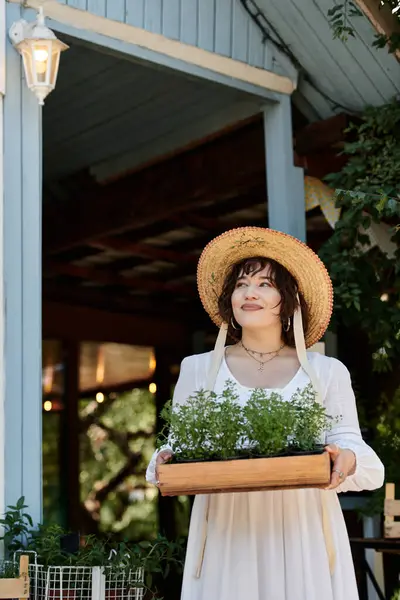 stock image A young woman in a white dress and straw hat stands in a summer garden, holding a tray of potted plants.