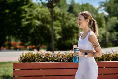 Spor kıyafetleri içinde vitiligo giymiş genç bir kadın elinde su şişesiyle bankın yanından geçerken gülümsüyor..