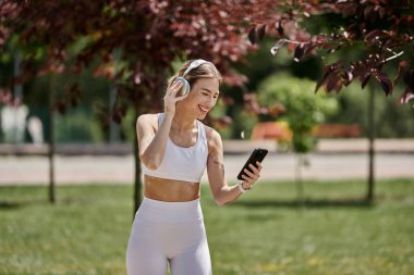 Beyazlar içindeki genç bir kadın müzik dinlerken gülümser ve parkta çalışırken telefonuna bakar..