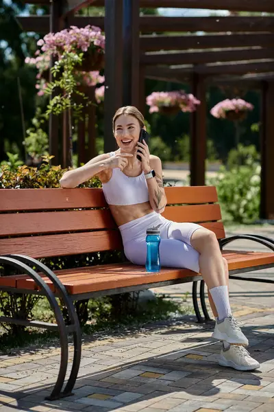 Stock image A young woman with vitiligo, wearing athletic clothes, sits on a park bench, laughing as she talks on her phone.