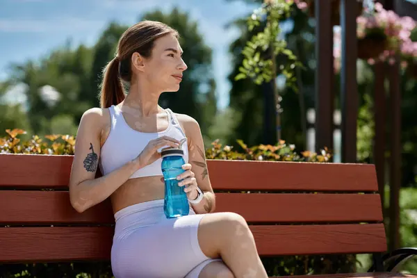 Stock image A young woman in activewear sits on a bench in a park, taking a break after a workout.