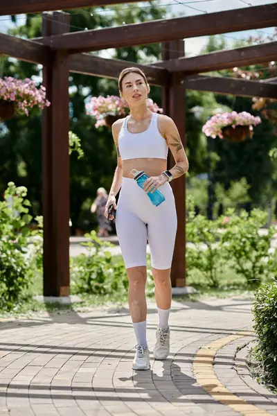 stock image A young woman in white athletic wear walks through a park with a water bottle in hand.