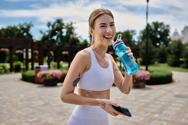 stock image A young woman in activewear smiles while drinking from a blue water bottle after a workout in a park.