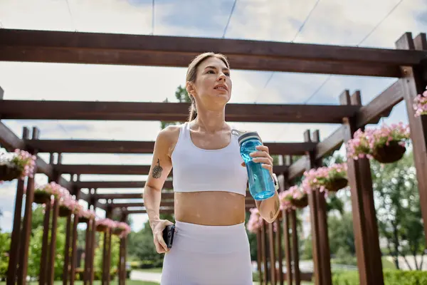 stock image A young woman with vitiligo, wearing white activewear, jogs through a park pergola, holding a blue water bottle.