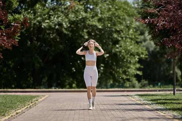 stock image A young woman with vitiligo in white activewear walks on a paved path in a park with trees and green grass on either side.