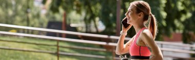 A young woman in a pink sports bra talks on the phone while taking a break from working out in a park. clipart