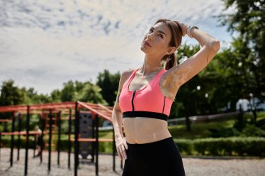Pembe ekili bluzlu genç bir kadın, sırtını açık havada, bulanık bir spor salonuna, gökyüzüne doğru çeviriyor..