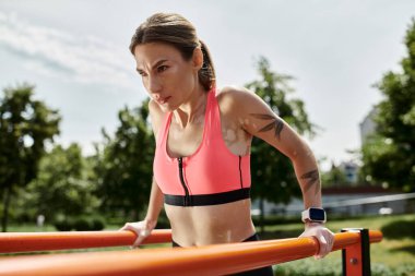 Pembe spor sutyenli genç bir kadın parktaki paralel çubuklarda çalışıyor, gücünü ve kararlılığını gösteriyor..