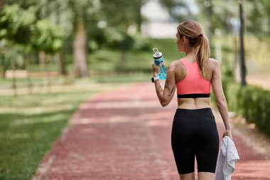 A young woman in activewear walks on a path in a park, holding a water bottle and a towel, after a workout. clipart