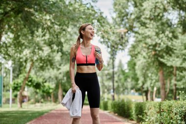 A young woman with vitiligo is walking through a park after a workout, smiling with a water bottle in her hand. clipart