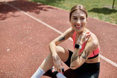 A young woman with vitiligo smiles while sitting on a red running track in a park. clipart