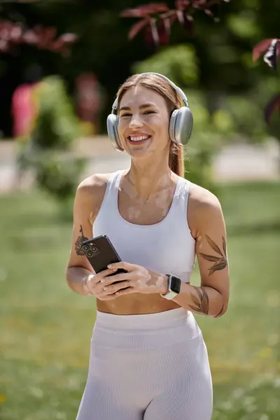 stock image A young woman in active wear, with vitiligo, walks in a park, holding her phone and smiling.