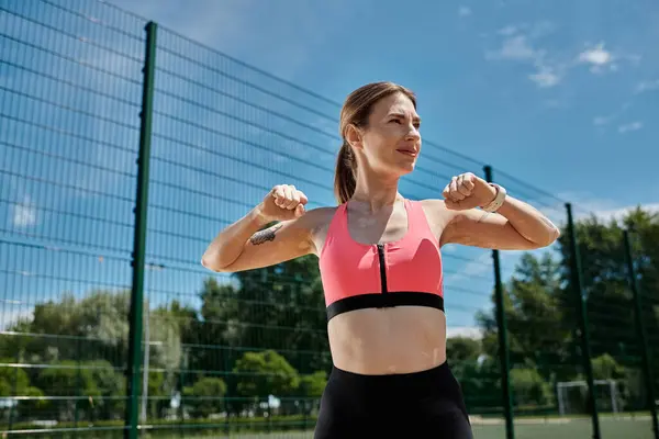 stock image A young woman in a pink sports bra and black leggings stretches her arms outdoors, against a green fence.