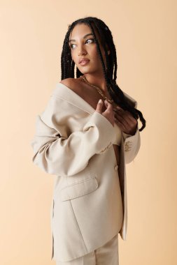 Örgülü genç siyah bir kadın, tarafsız bir arkaplanda bej bir ceketle poz veriyor..