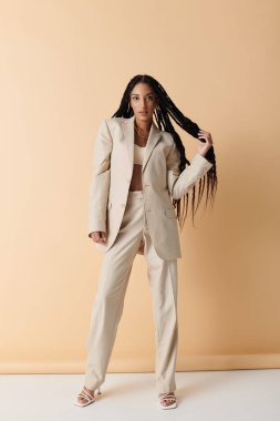 Uzun örgülü siyah bir kadın bej bir takım elbiseyle poz veriyor, eli saçında dinleniyor..