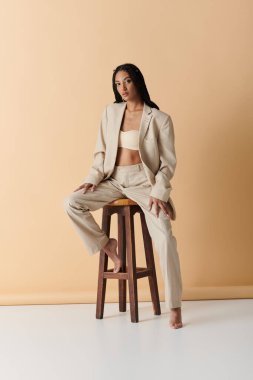 Bej takım elbiseli genç bir kadın tahta bir taburede soluk bej bir zemine karşı oturuyor..