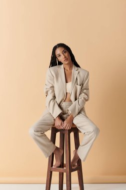 A young woman sits on a wooden stool, dressed in a beige suit, her legs crossed and her arms resting on her knees. clipart