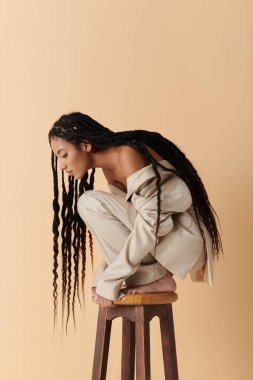 Beyaz ceketli, 20 'li yaşlarda şık Afro-Amerikan bir kadın, bej arka planda uzun örgülü, tahta bir taburede oturuyor.