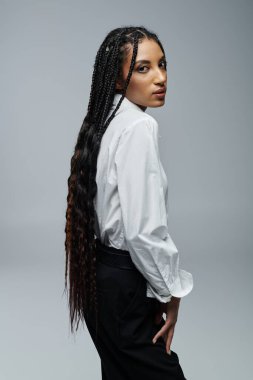 Uzun örgülü şık bir genç kadın beyaz bir gömlekle gri bir fonda poz veriyor..