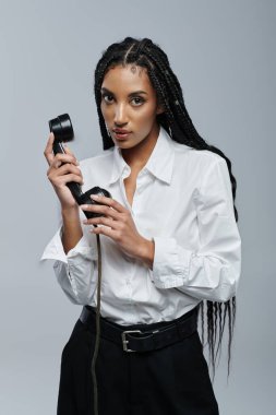 Afrika kökenli Amerikalı bir kadın eski moda bir telefon tutuyor. Gri bir fon karşısında izleyiciye bakarken gizem saçıyor.