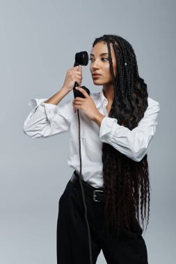 A young woman with long braids poses in a white shirt against a grey background, holding a vintage phone to her ear. clipart