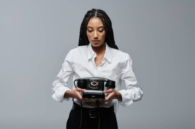 A young woman with braids in a white shirt holds a vintage black rotary phone, looking down at it with an air of contemplation. clipart