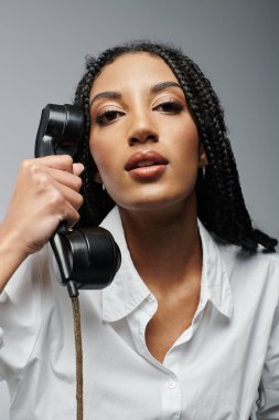 Saçları örgülü bir kadın ahizeyi yakın tutarak bir telefon konuşmasını dinliyor. Gri bir arka plana karşı beyaz bir gömlek giyiyor.