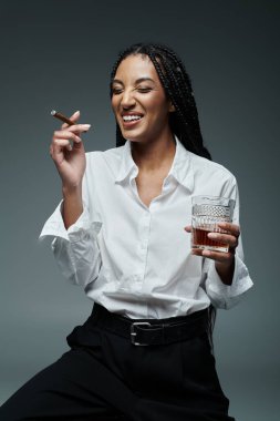 A stylish woman wearing a white shirt and black pants laughs while holding a cigar in one hand and a drink in the other. clipart