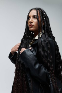 A young woman with long braids and a stylish black leather jacket poses against a grey backdrop, radiating confidence and individuality. clipart