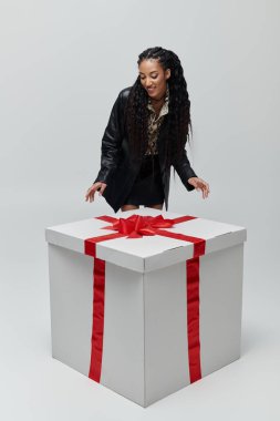 A stylish African American woman in a black leather jacket and animal print shirt leans over a large, white gift box with a red bow. clipart