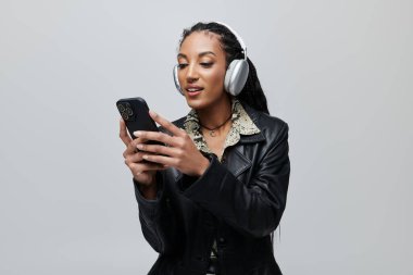 A young woman with long black braids wears a black leather jacket over an animal print shirt, headphones, and is using her phone. clipart