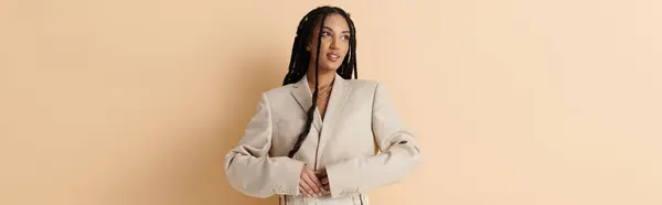 stock image A young woman with long braids smiles while wearing a chic beige blazer against a neutral backdrop.