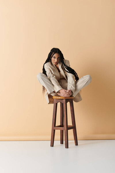 Молодая женщина в светлом костюме сидит скрестив ноги на деревянном стуле на бежевом фоне.