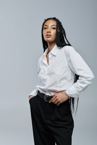A young woman in a white shirt and black pants poses against a grey backdrop. She has black braided hair and a confident expression.