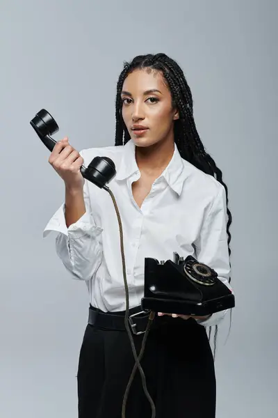 stock image A young woman with braided hair poses with a vintage black phone in her hand. She is wearing a white button-down shirt and black pants.