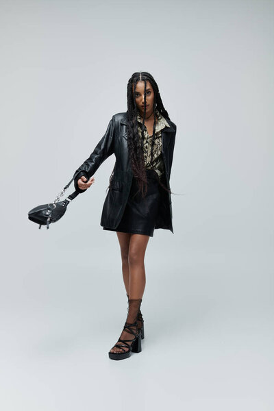 Уверенная молодая женщина в черной кожаной одежде с рубашкой с отпечатком животного и высокими каблуками позирует на сером фоне