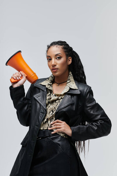 Молодая женщина с косичками позирует в черной кожаной куртке и юбке, держа в руках оранжевый мегафон.