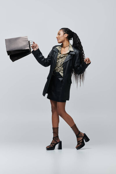Молодая женщина в черной кожаной куртке и юбке позирует с сумками в руке.