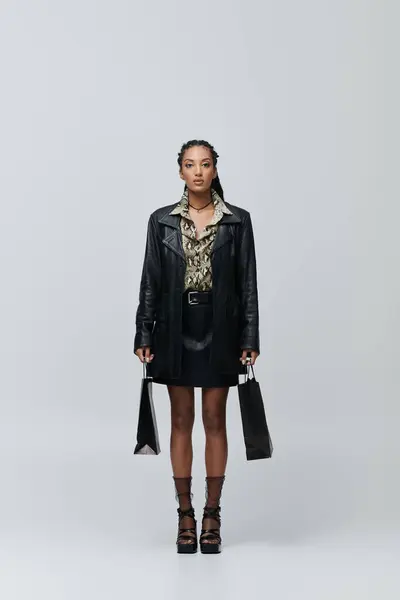 stock image A stylish young woman poses in a black leather jacket and skirt, with a patterned shirt, holding shopping bags.