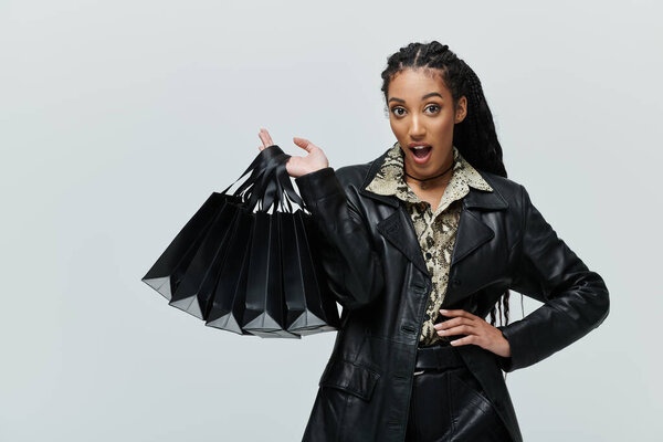 Молодая женщина в черной кожаной куртке и рубашке животного принта держит стопку сумок с покупками, выглядящих удивленными и восхищенными.