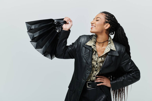 Молодая женщина в черной кожаной куртке и рубашке со змеиным отпечатком держит стопку сумок для покупок, счастливо улыбаясь.