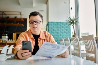 A man with Down Syndrome sits at a cafe table looking at a menu and using his phone. clipart