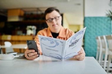 Down sendromlu, gözlüklü ve turuncu gömlekli bir adam elinde akıllı telefon ve menü olan bir kafede menüye bakıyor.