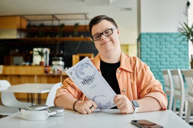 Down sendromlu bir adam bir kafe masasında oturmuş, menüyü tutarken gülümsüyor ve yemeğini seçiyor..