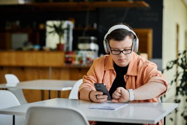 Down sendromlu bir adam kafe masasında oturup telefonundaki menüye bakıyor. Kulaklık takıyor ve akıllı kaset takıyor..